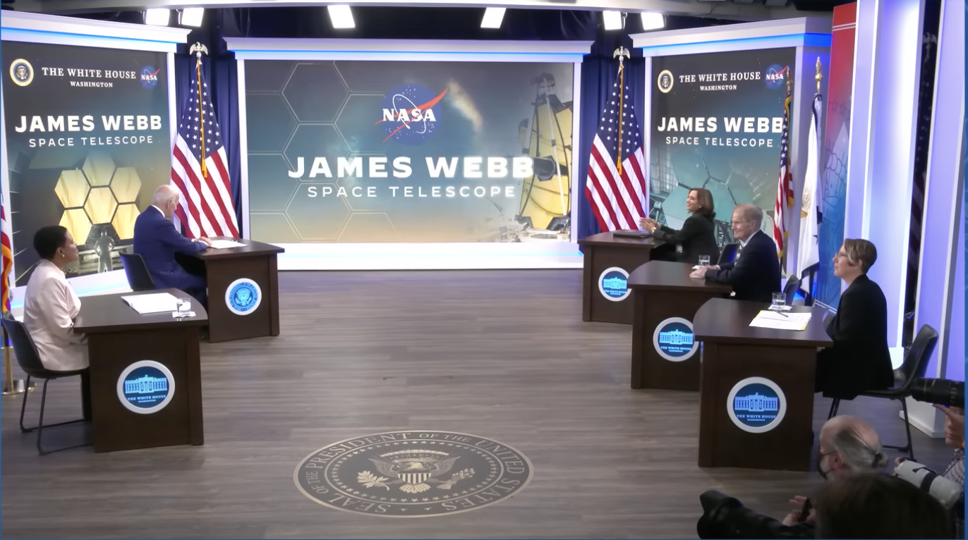 President Biden and other VIPs sit at small desks in front of a screen saying "James Webb Space Telescope" for the White House briefing event.